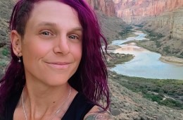 A person with shoulder-length dyed-purple hair looks into the camera for a selfie in front of a river that runs through the base of a canyon.