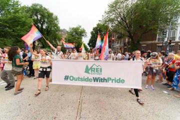A group of people march in a Pride parade, holding an REI Co-op Outside with Pride banner.