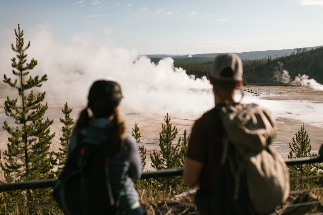 Two hikers wearing packs look at a geyser