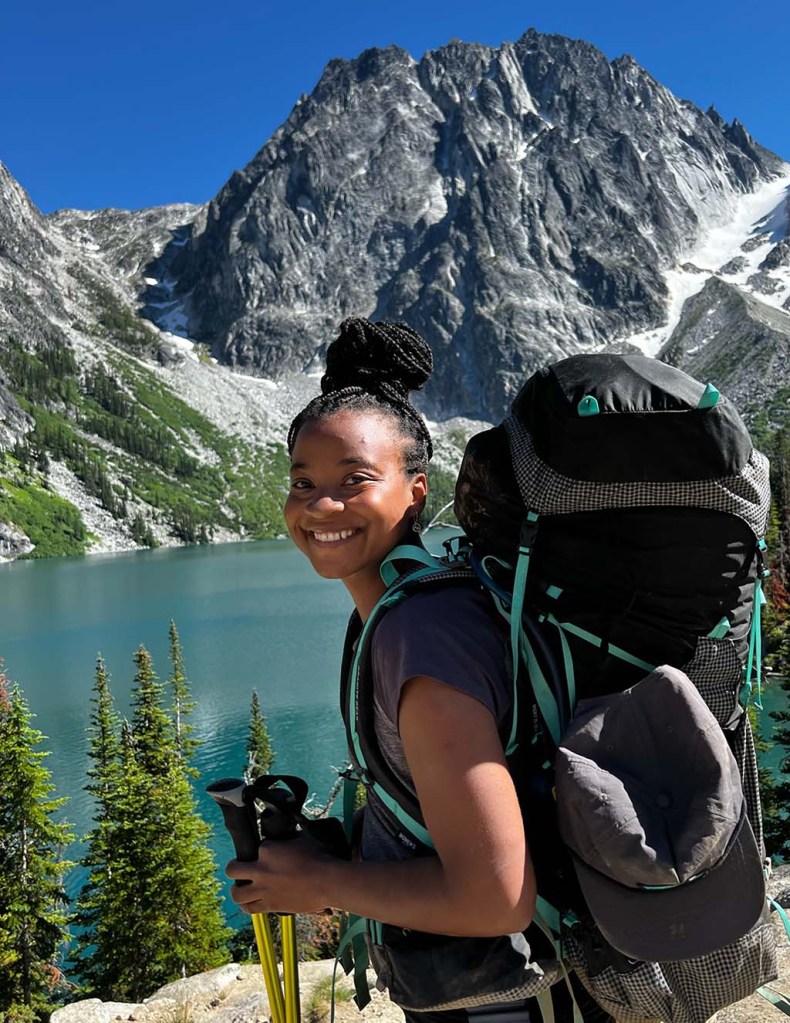 A person with a large pack on smiles for the camera in front of a glacial lake and a mountain peak