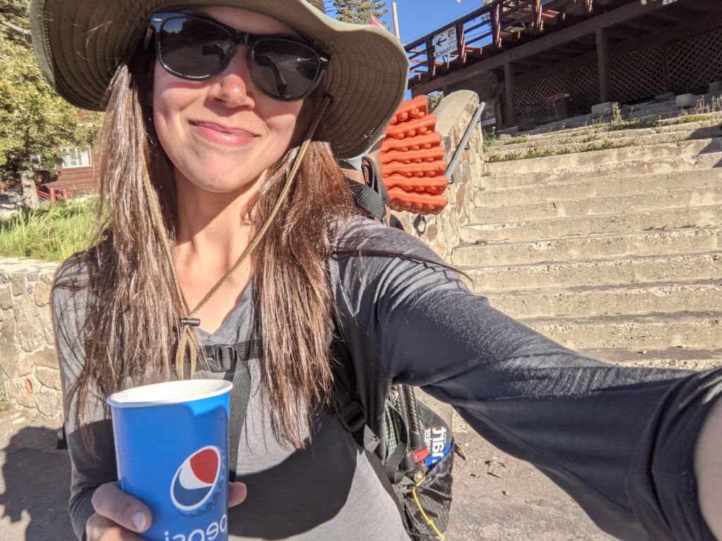 A white person with long blondish hair wearing sunglasses and a floppy hat smiles into the camera during a selfie.