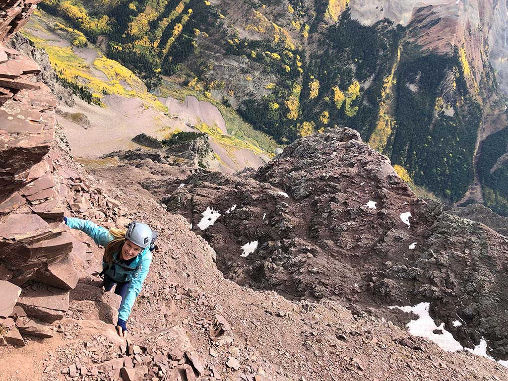 A person in a teal blue top and climbing helmet scales a mountain wall.