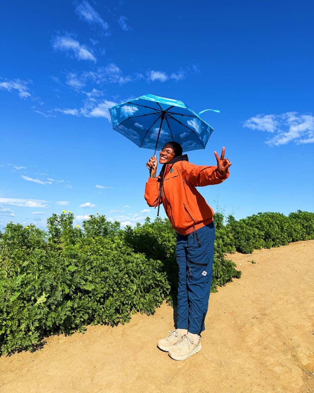 A person wearing a bright orange top and blue pants smiles and holds an umbrella with a blue sky and cloud design on it.