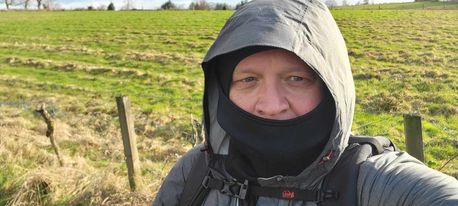 A man wearing a black hood and black mask stands in front of a field