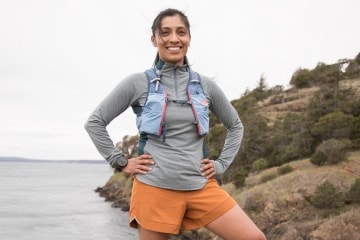 A person standing near a cliff, smiling, in running shorts, a shirt and a running vest.