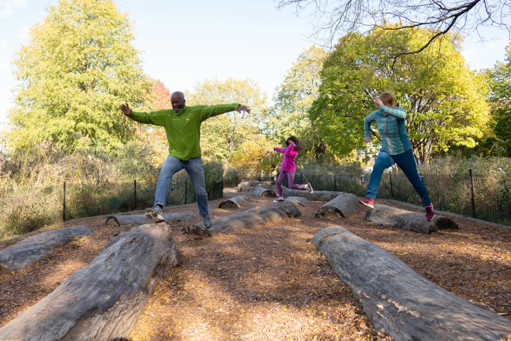 A family of three is playing in a park with a feature that includes laid-down logs to climb and jump over.