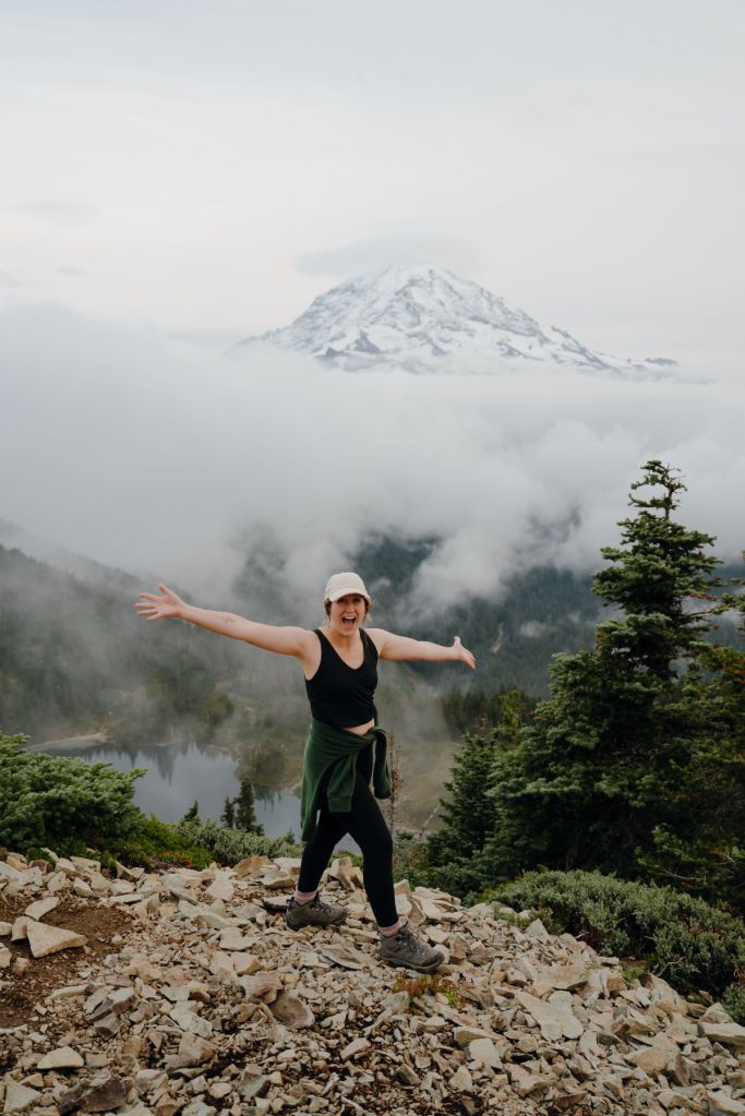 A hiker with a wide smile stands with arms outstretched at the top of a mountain.