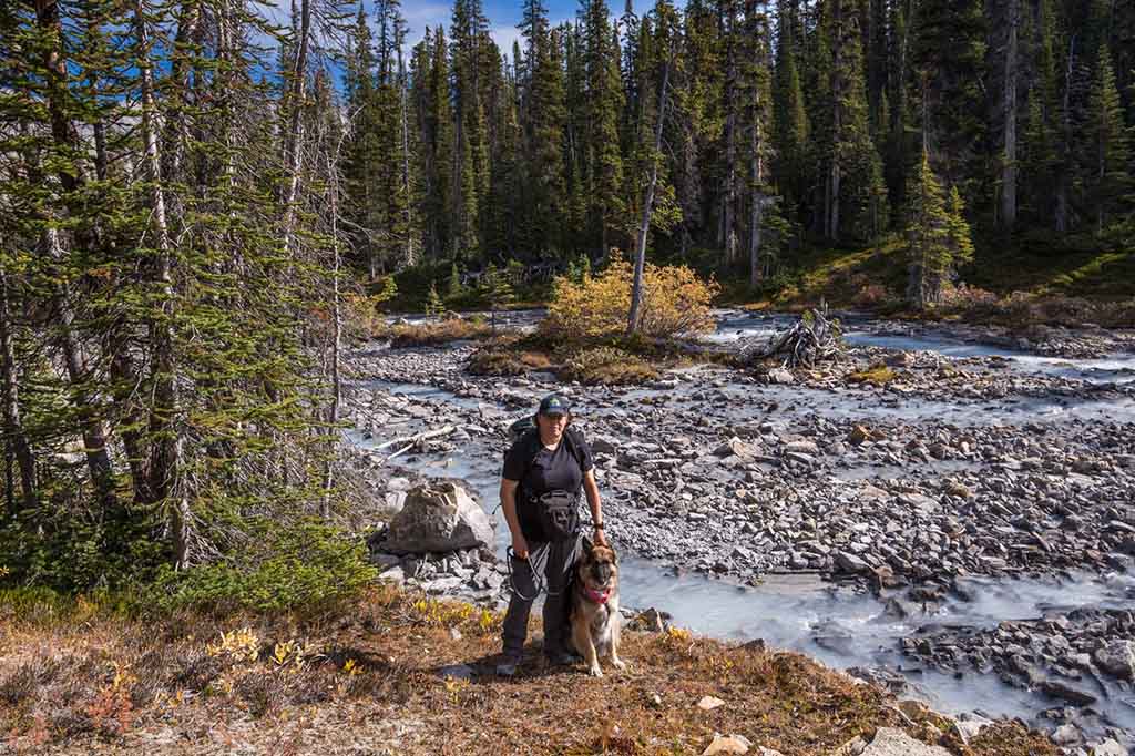 A hiker wearing a packpack stands next to a glacial river with their dog