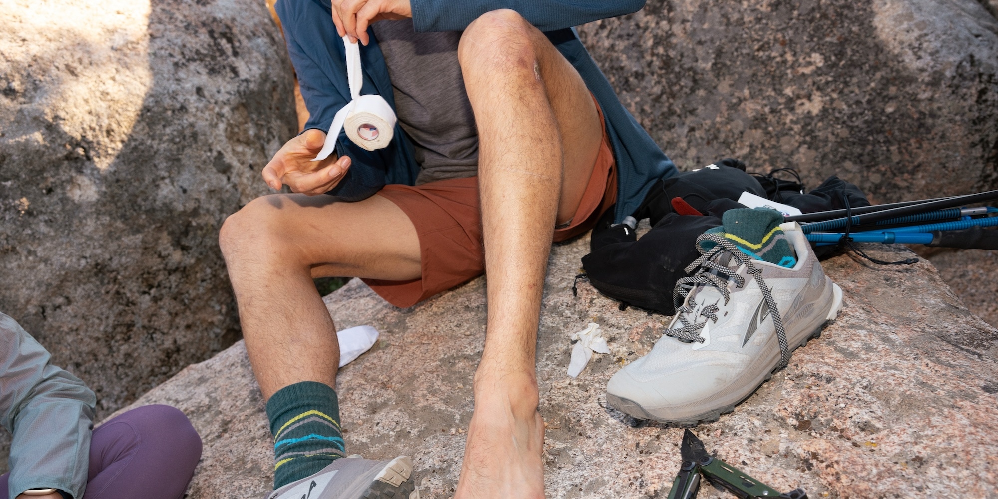 A runner sits on a rock beside a trail and applies adhesive fabric tape.