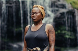 A Black woman wearing glasses, a headwrap and a tank top, standing in front of a waterfall.