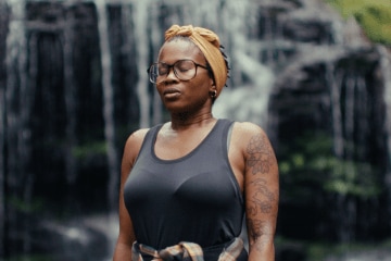 A Black woman wearing glasses, a headwrap and a tank top, standing in front of a waterfall.