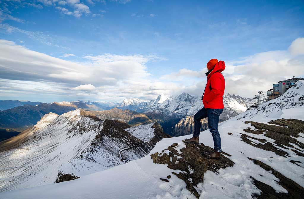 A person in denim pants and a bright red jacket stands at the top of a mountain