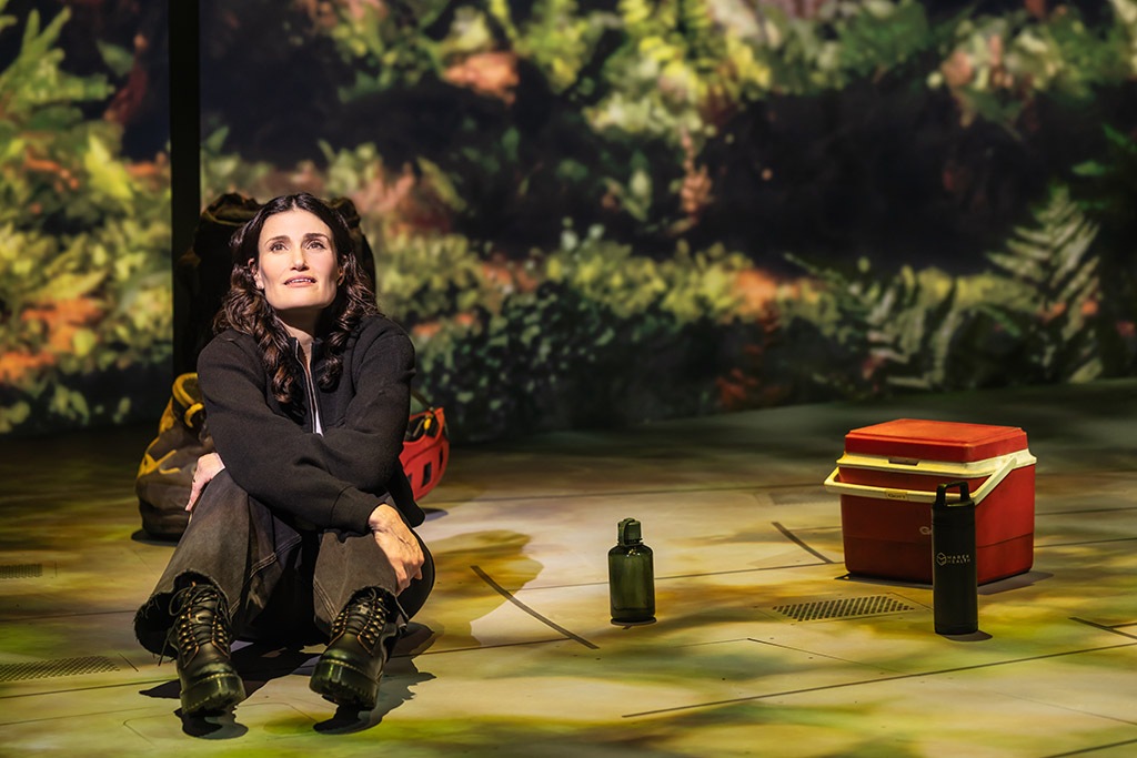 A person sitting on a stage next to a water bottle and a cooler, with a forest backdrop behind her.