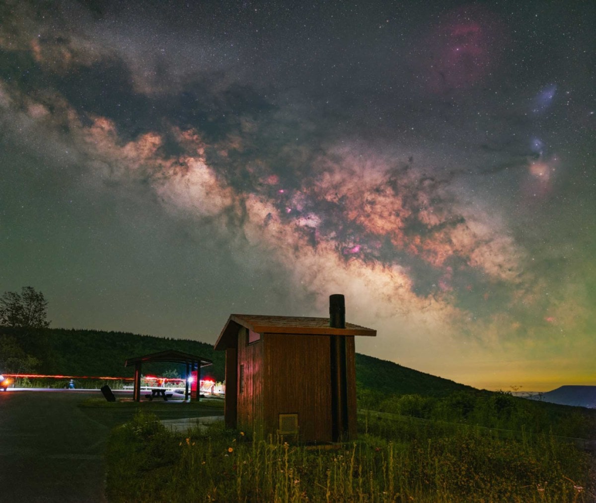 A small cabin in the foreground is set against a backdrop of the Milky Way