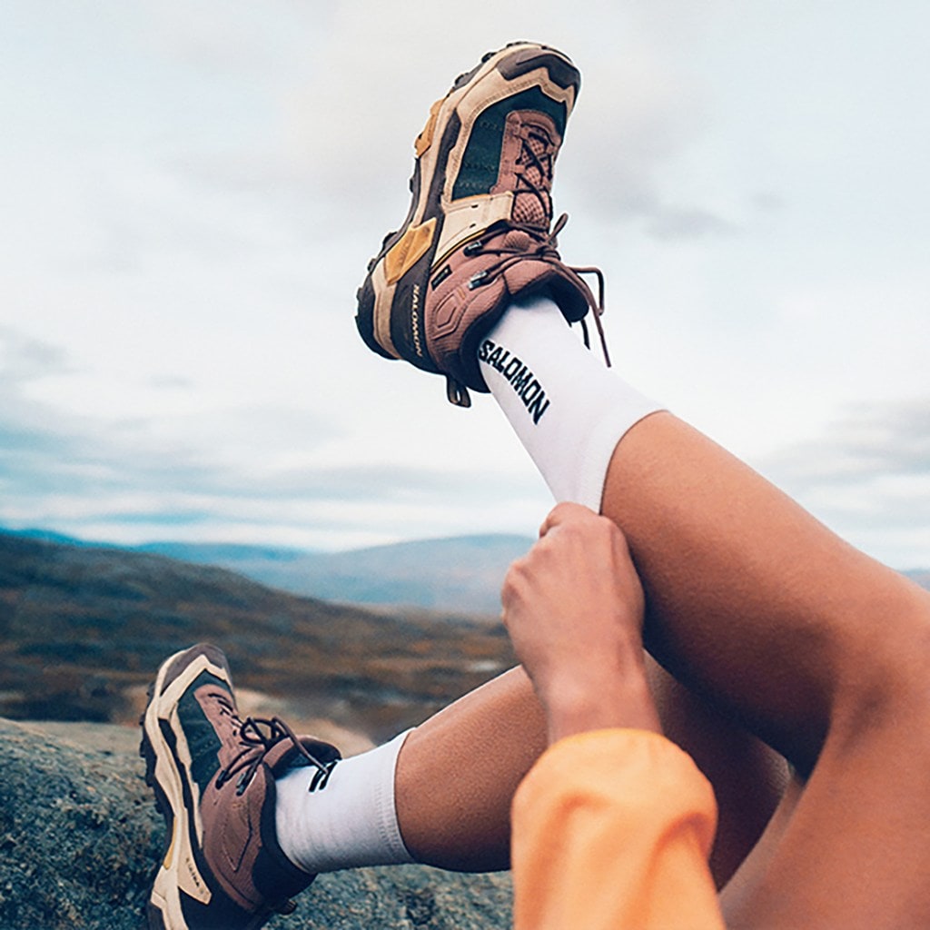 A person leaning back raises their left leg in the air to pull up a pair of socks that say Salomon; they are wearing mid-height hiking boots.