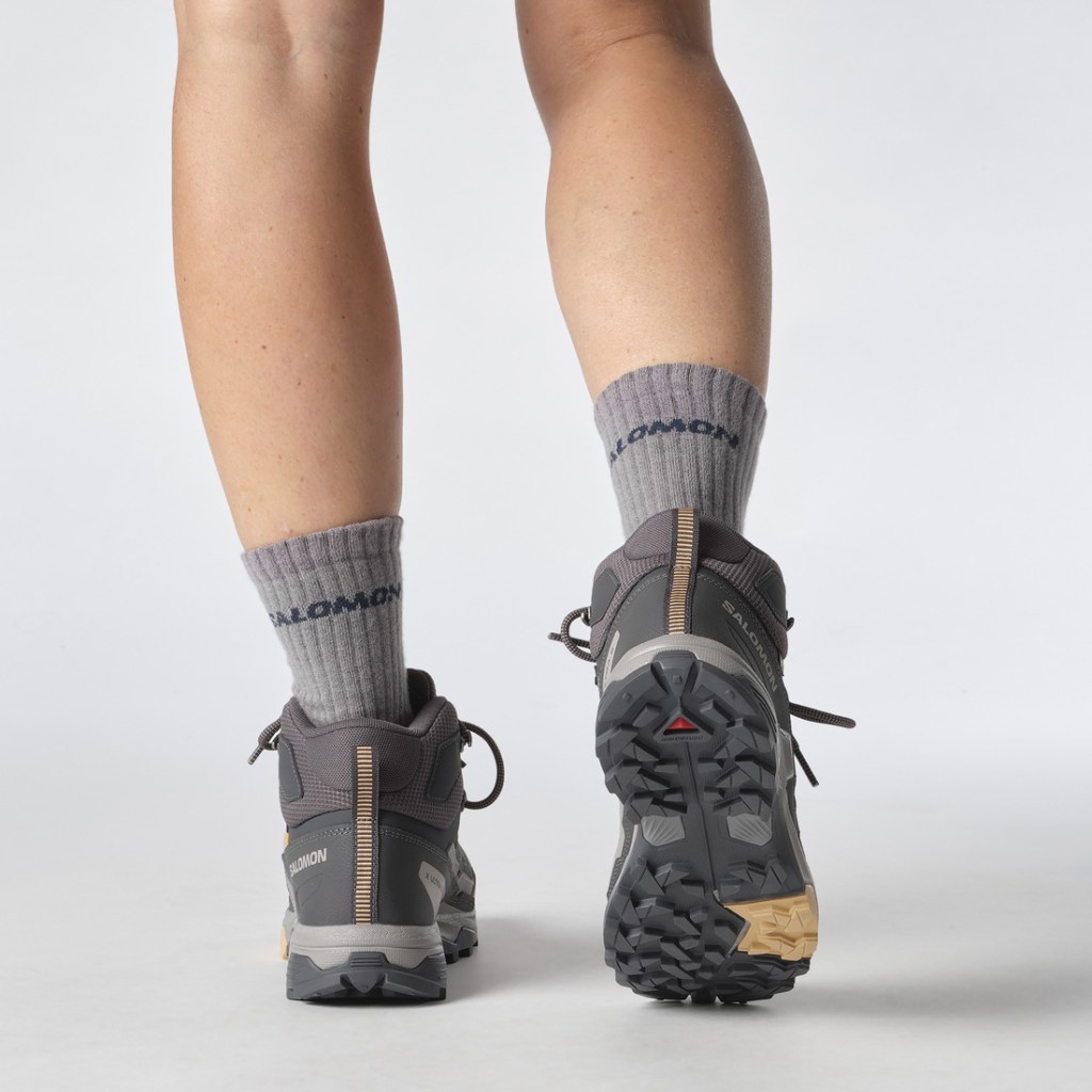 A person wearing shorts standing facing away from the camera is shown from the knee down, displaying the tread on the Salomon X Ultra 5 Mid GORE-TEX Hiking Boots.