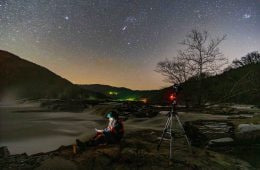 A person sits by a body of water, illuminated by his cell phone light. Next to him is a camera, and above him the sky is full of stars