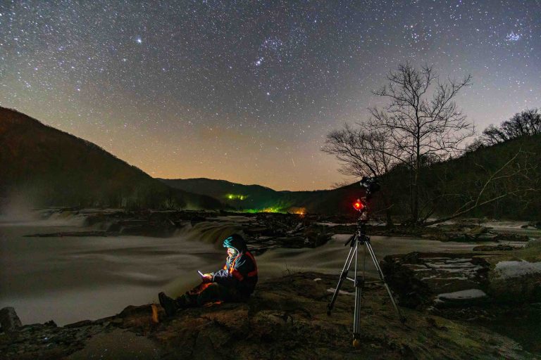 A person sits by a body of water, illuminated by his cell phone light. Next to him is a camera, and above him the sky is full of stars