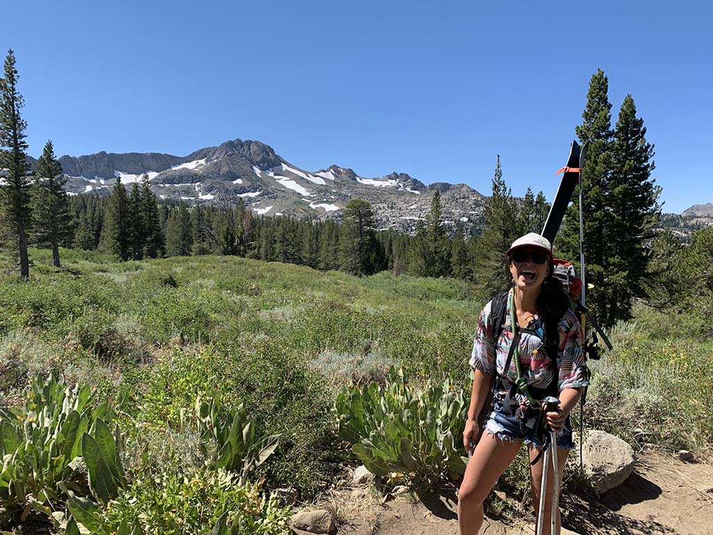 A person in a hat and sunglasses takes a selfie at the top of a mountain ridge