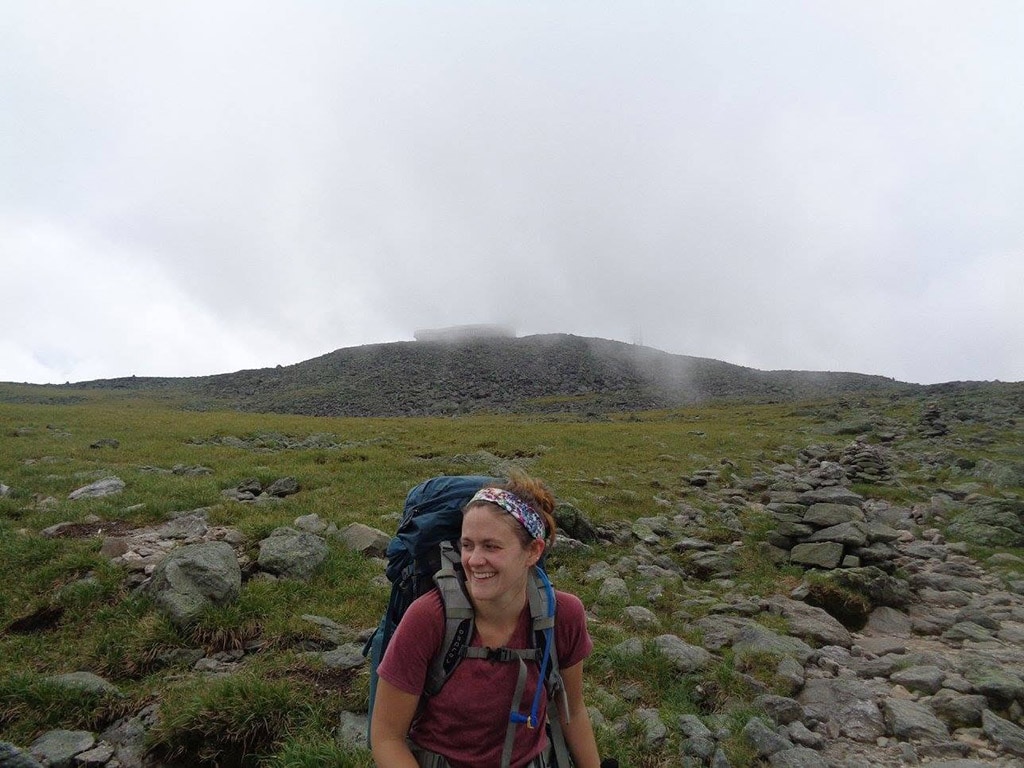 A hiker stands in the foreground, in front of a mossy, mist-obscured hill.