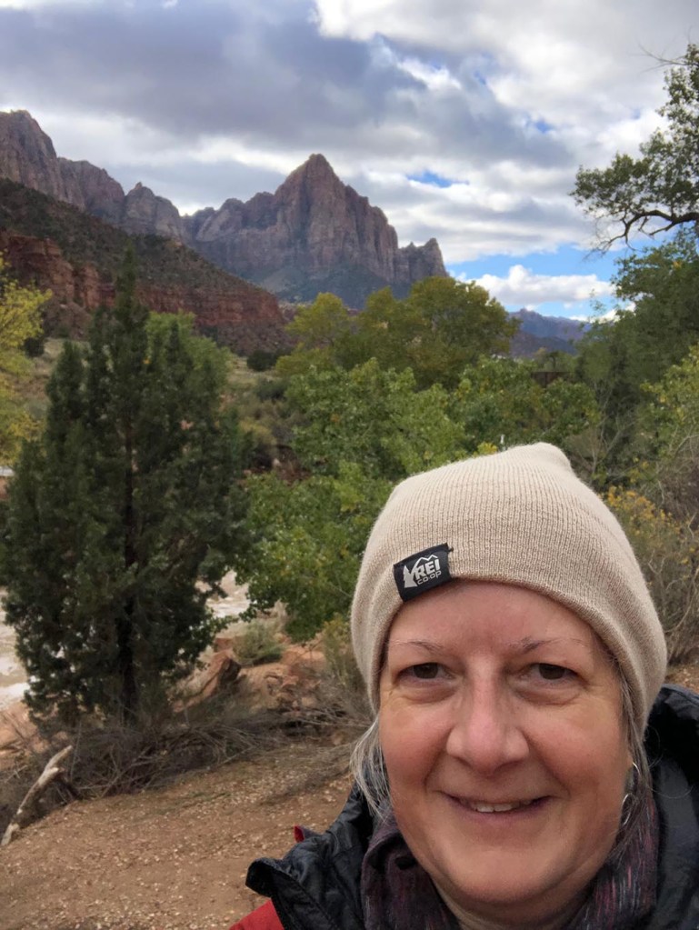 A person in a beige knit hat takes a selfie in front of a beautiful mountain landscape