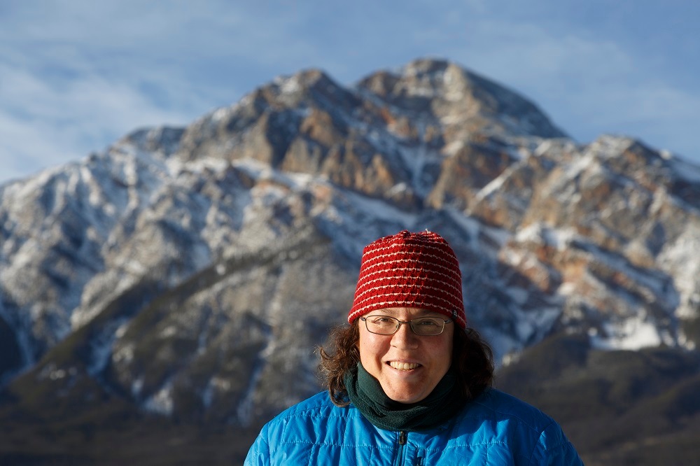 A person standing in front of a mountain peak, smiling, dressed in a hat and jacket