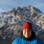A person standing in front of a mountain peak, smiling, dressed in a hat and jacket