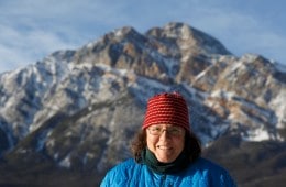 A person standing in front of a mountain peak, smiling, dressed in a hat and jacket