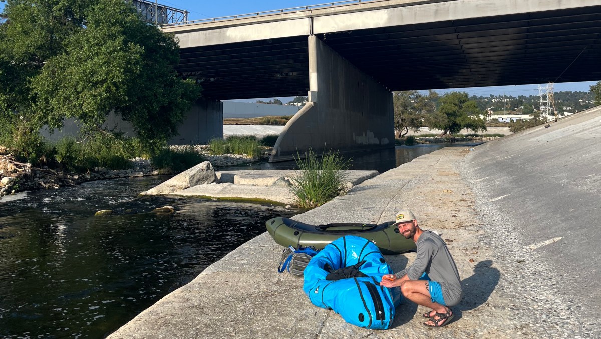 A person crouches by the side of the LA river, near an overpass, setting up a packraft.