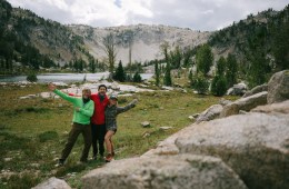 Three people wave at the camera against a mountain backdrop