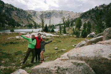 Three people wave at the camera against a mountain backdrop