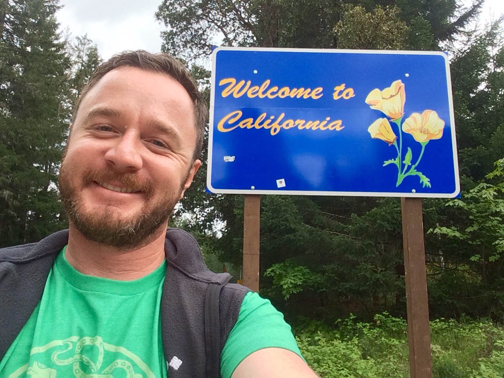 A smiling person poses in front of a Welcome to California sign