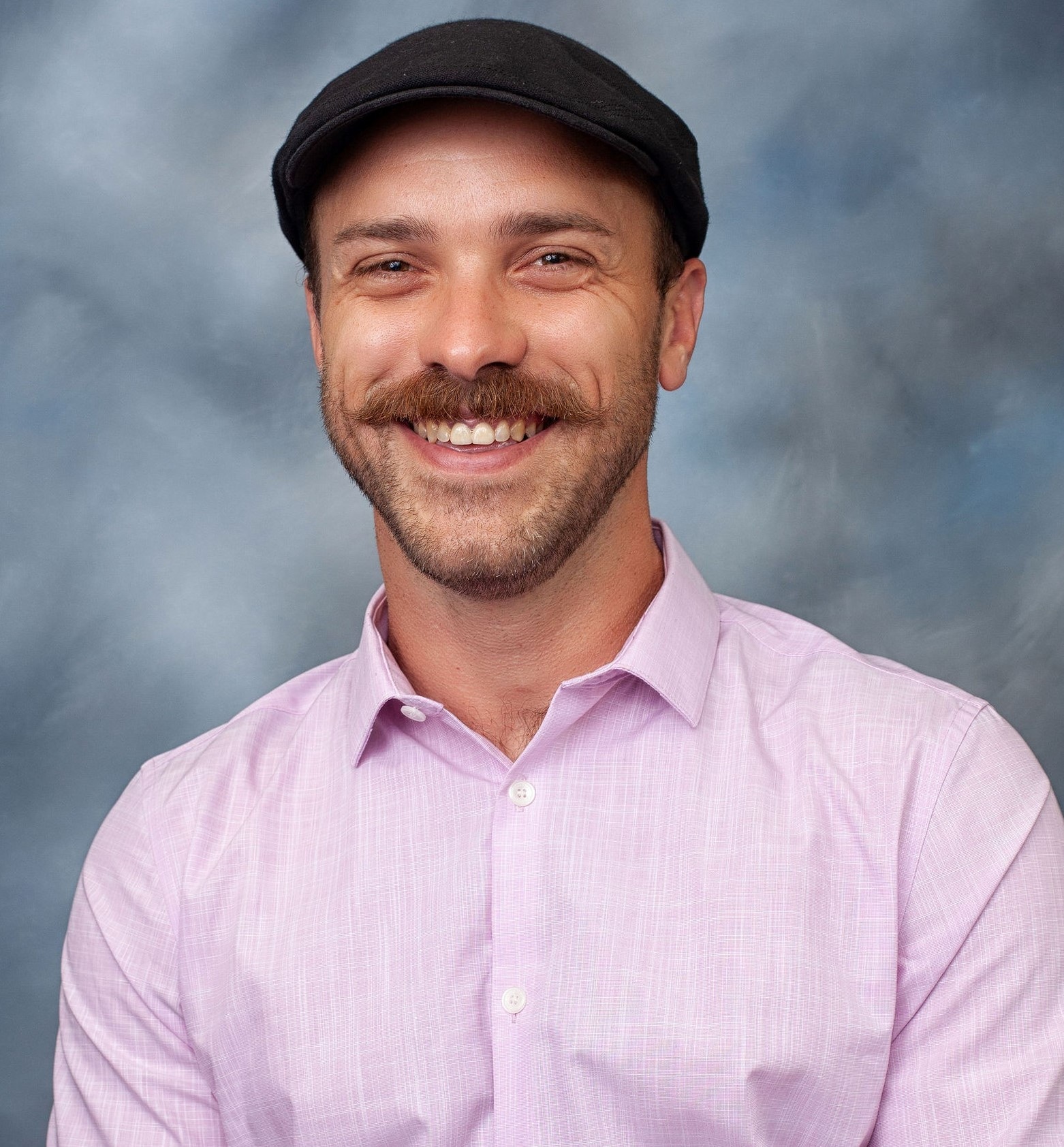 A studio portrait of a person with a moustache and a newsboy cap