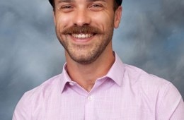 A studio portrait of a person with a moustache and a newsboy cap