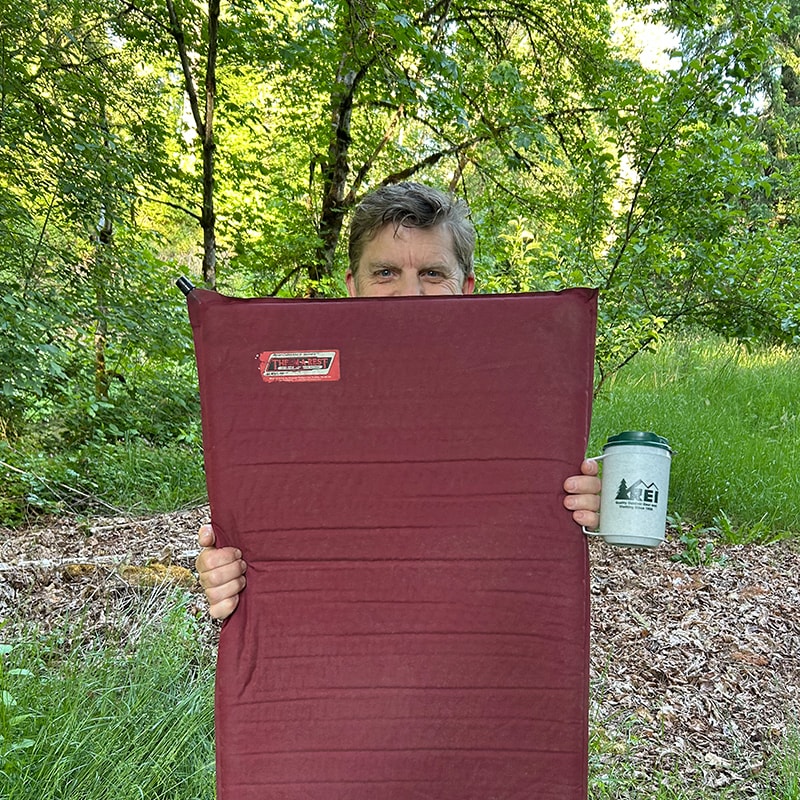 Greg Wayt holds a maroon sleeping pad and classic REI insulated mug
