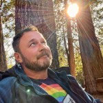 A person wearing a rainbow shirt sits among the trees, looking up at the canopy