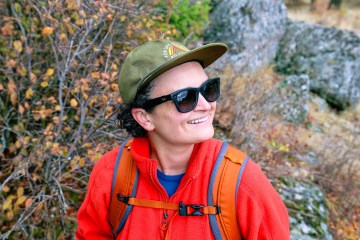 A person wearing an REI Co-op hat, orange fleece jacket and Ray-Ban sunglasses smiles off into the distance.