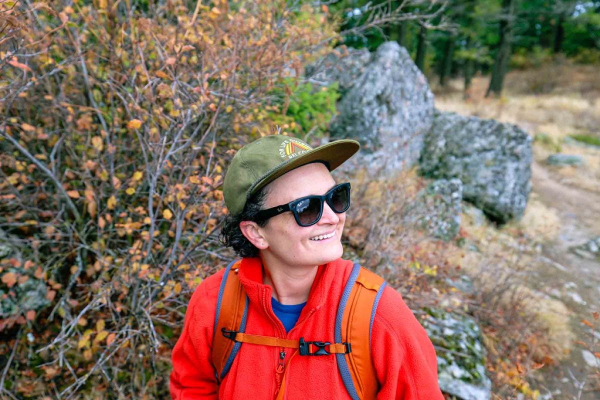 A person wearing an REI Co-op hat, orange fleece jacket and Ray-Ban sunglasses smiles off into the distance.