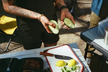 A person at a campsite slices avocado and citrus to cook a meal.