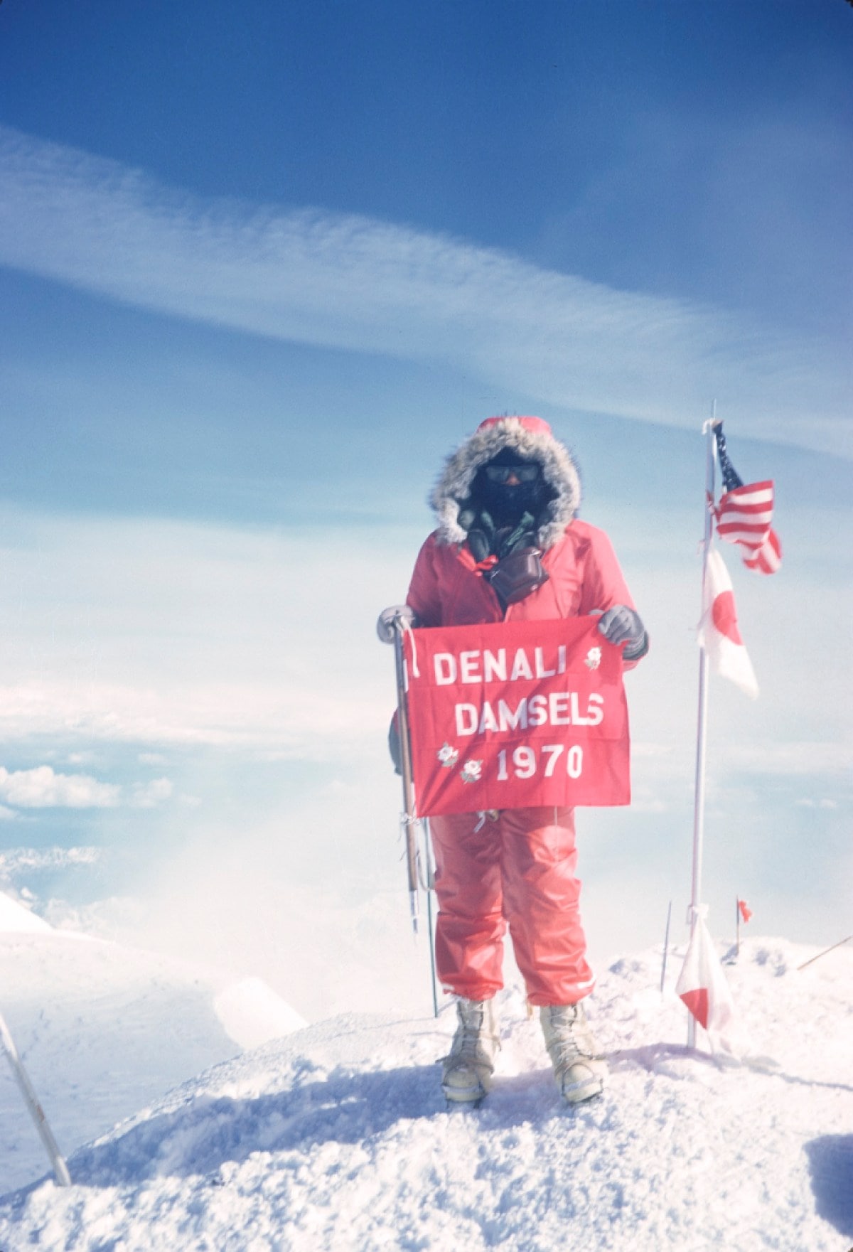 A climber in full gear stands at a mountain summit next to an American flag holding a sign that reads Denali Damsels