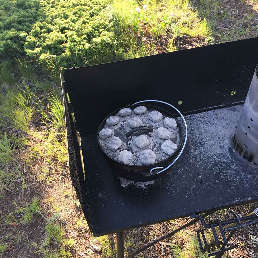 A cast iron Dutch oven with a lid sits on a metal table; there are several white-hot charcoal briquettes on the pot's lid
