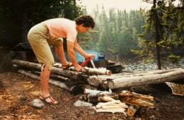 A person in the woods is bent over a downed tree and uses a portable, folding saw.