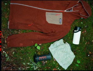 A Patagonia fleece, an AeroPress coffee maker, and a thermos are laid down on the grass.