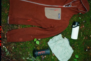 A Patagonia fleece, an AeroPress coffee maker, and a thermos are laid down on the grass.