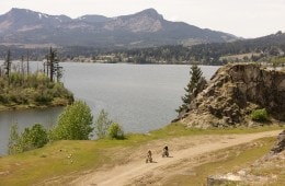 Two people riding bikes along a dirt trail. A large body of water is in the distance.
