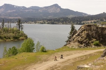Two people riding bikes along a dirt trail. A large body of water is in the distance.