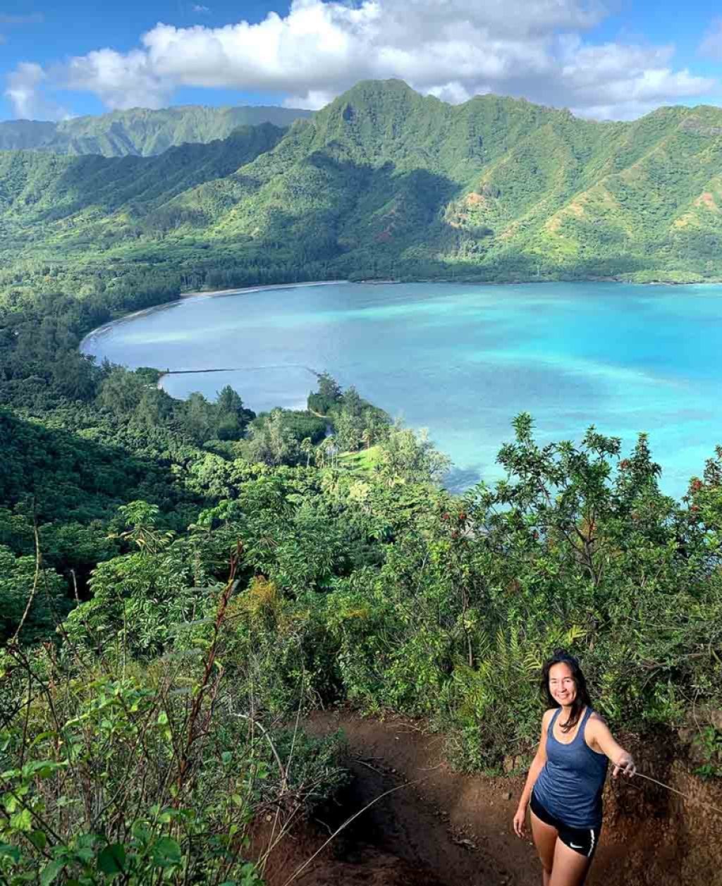 A person stands in the foreground, with a clean blue body of water behind them, with mountains in the background.