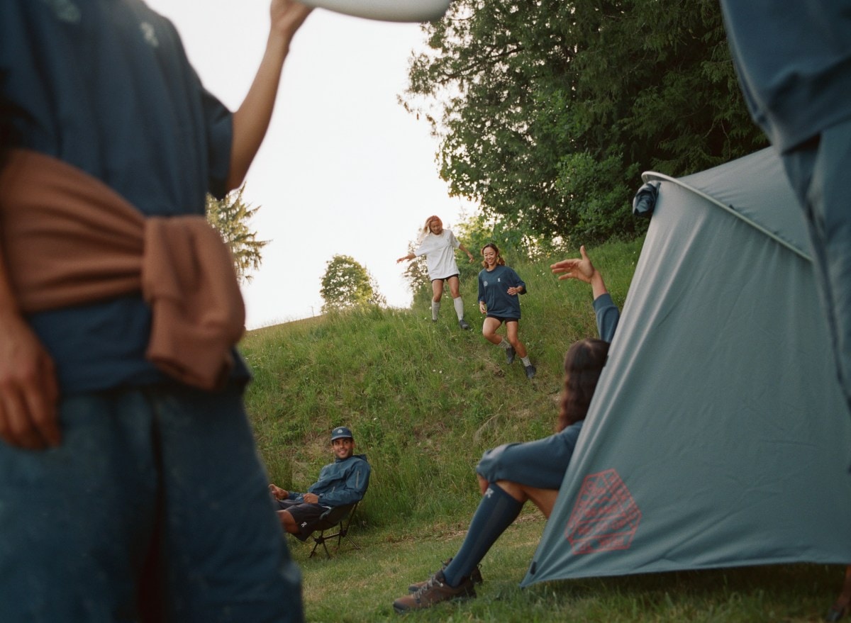 A group of people gather at a campsite with a tent in the foreground; several folks are seen running down a grassy hill.