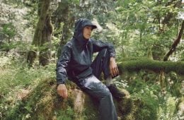 A man in navy-colored rainwear and trail-running shoes sits on a mossy rock in a lush forest.