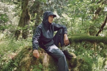 A man in navy-colored rainwear and trail-running shoes sits on a mossy rock in a lush forest.